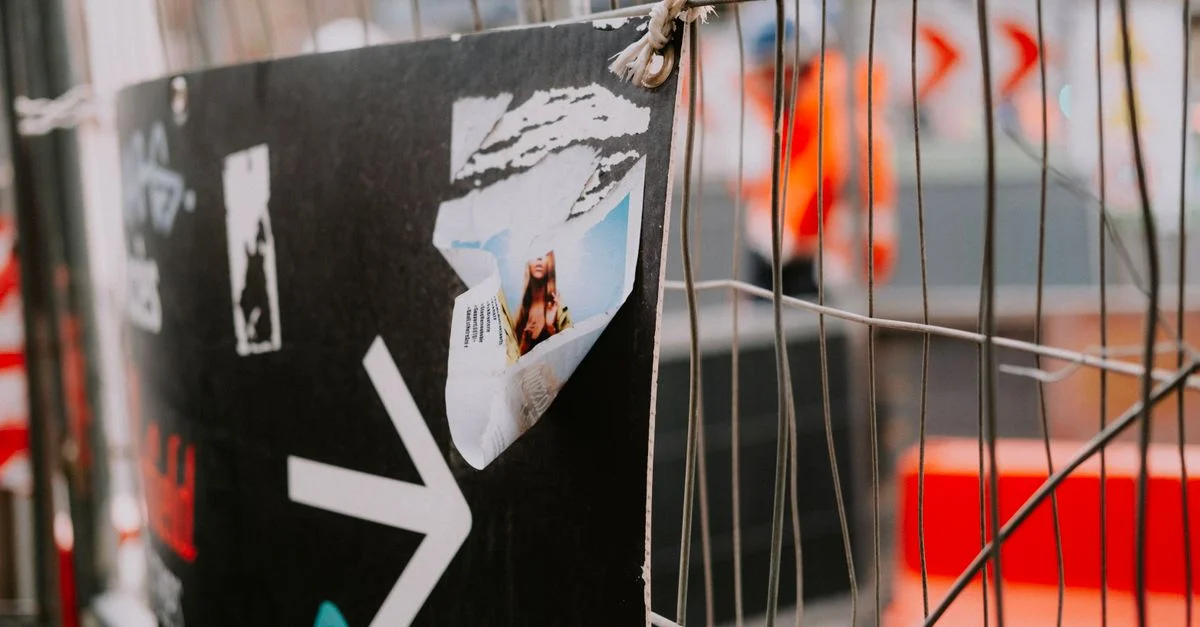 Une affiche déchirée représentant une personne est accrochée à une clôture en métal, avec un arrière-plan flou, en extérieur.