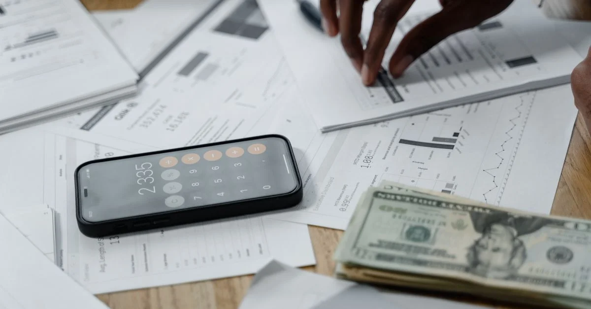 Gros plan sur des mains calculant des données financières avec des rapports, un smartphone et des espèces sur la table.