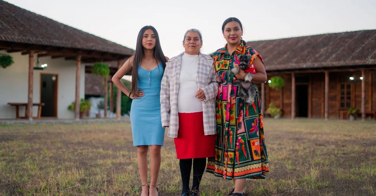 Trois femmes se tiennent ensemble devant une maison rustique traditionnelle, présentant des tenues culturelles diverses.