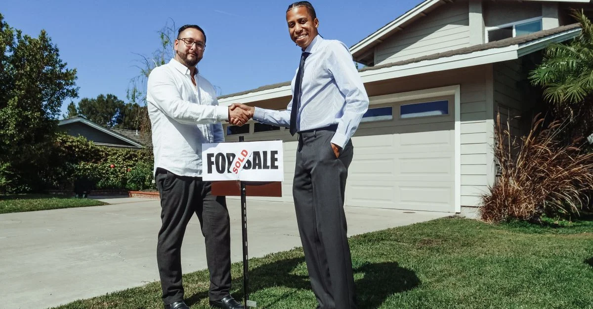 Deux hommes se serrent la main devant une nouvelle maison avec un panneau 'vendu', symbolisant un accord immobilier réussi.