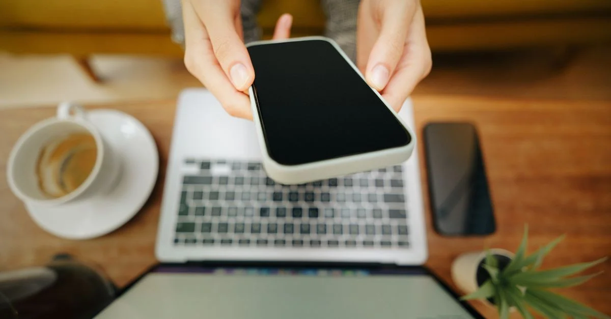 Mains tenant un smartphone sur un ordinateur portable dans un bureau à domicile confortable avec café et plante.