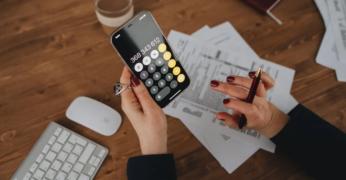 Femme calculant ses dépenses avec une calculatrice smartphone sur un bureau en bois avec des documents.